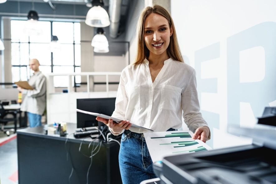 A smiling woman in a white shirt and jeans holds papers near a printer in a modern office.