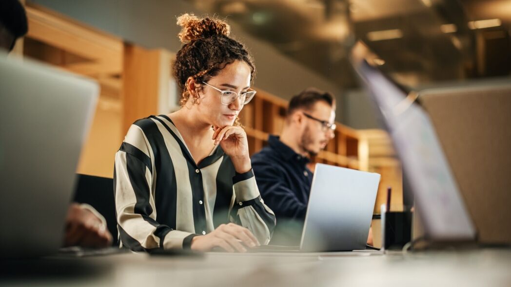 A woman with curly hair and glasses, wearing a striped shirt, works on her laptop in a modern office setting.