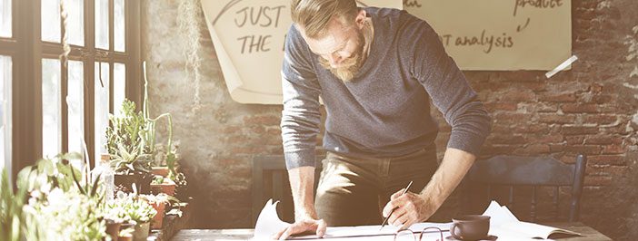 A bearded man in a casual sweater leans over a desk, writing on a document in a sunlit room filled with plants.
