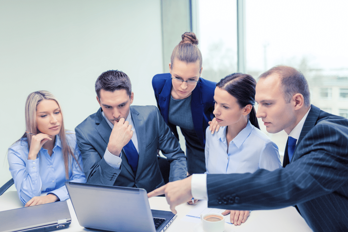 A group of five business professionals are gathered around a laptop, engaged in a discussion.