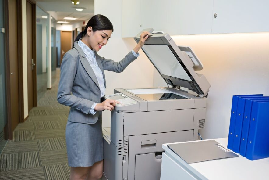 A smiling woman in a business suit operates a large office copier, with its lid open, in a well-lit office environment.