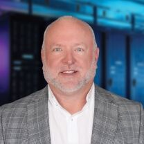 A smiling man with a beard and a grey plaid jacket stands in front of a blurred background of server racks.