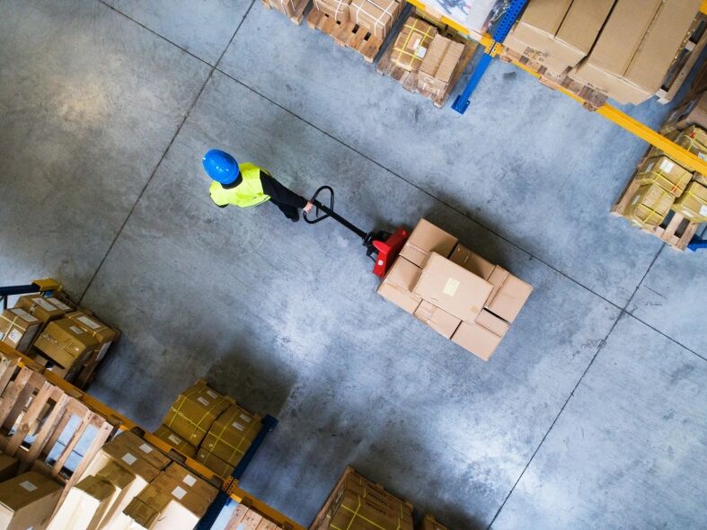 A worker in a high-visibility vest and hard hat operates a pallet jack loaded with boxes in a warehouse.