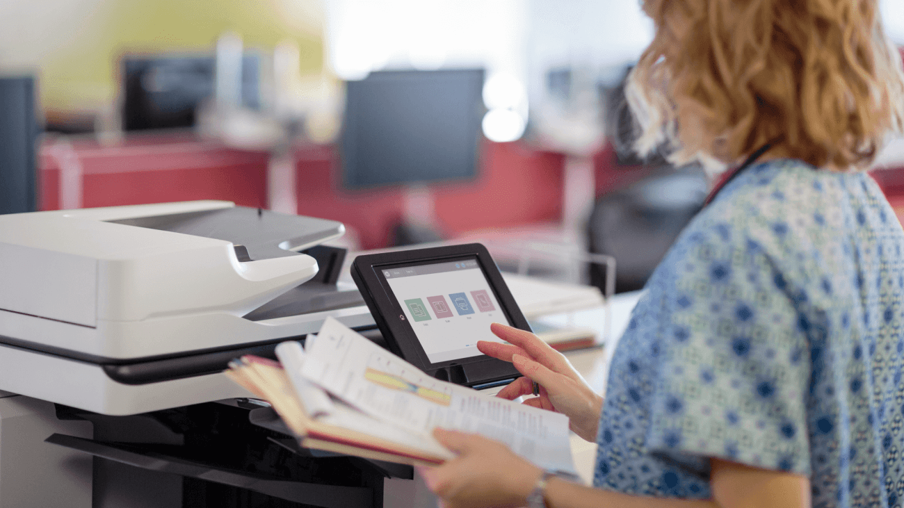 A person in a blue patterned shirt uses a touchscreen interface on a scanner to operate the device, holding a stack of