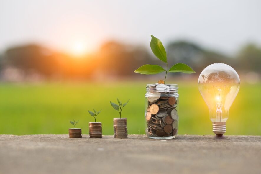 A glowing lightbulb and a jar filled with coins, with stacks of coins topped by small plants, symbolize financial