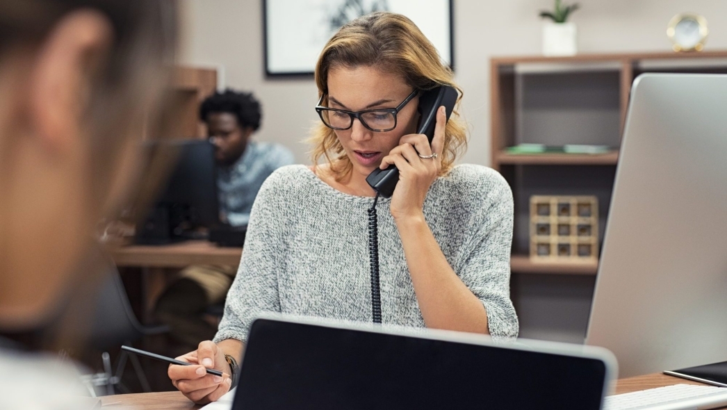 A woman in glasses talks on a corded phone while holding a pencil and looking at a laptop.