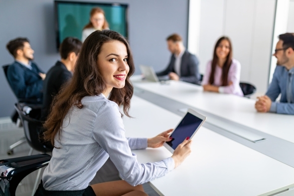 A young woman smiles at the camera while holding a tablet in a business meeting with colleagues in the background.