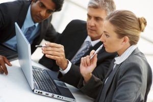 Three professionals in business attire collaborate around a laptop, engaged in a discussion or presentation.